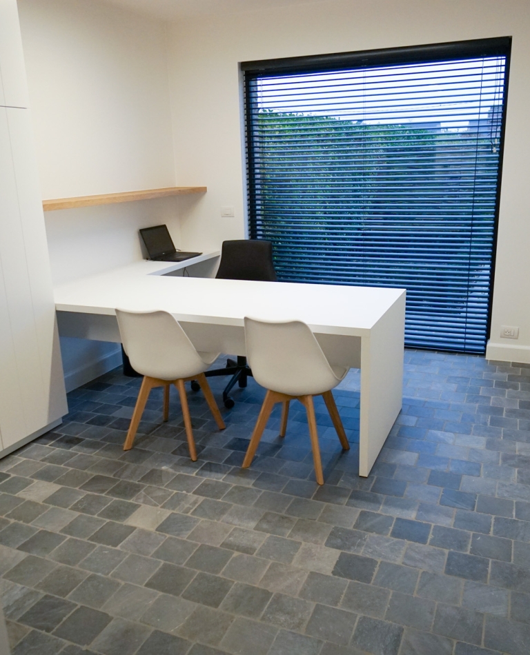 nterieur op maat voor een logopediepraktijk in Oostnieuwkerke met een volledig op maat gemaakte kastenwand, inclusief boven- en onderkasten met verborgen scharnieren. Strakke afwerking in wit, gecombineerd met een groot bureau en raam met zwarte lamellen dat natuurlijk licht binnenlaat