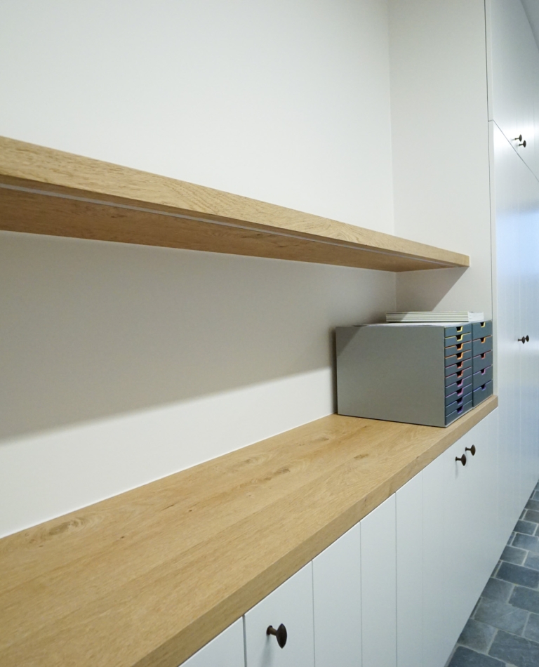 Detail van een maatwerk interieur in Oostnieuwkerke met op maat gemaakte inbouwkasten en een doorlopende houten werkblad- en wandplank. Strakke witte kasten met donkere knoppen, gecombineerd met een modern bureau