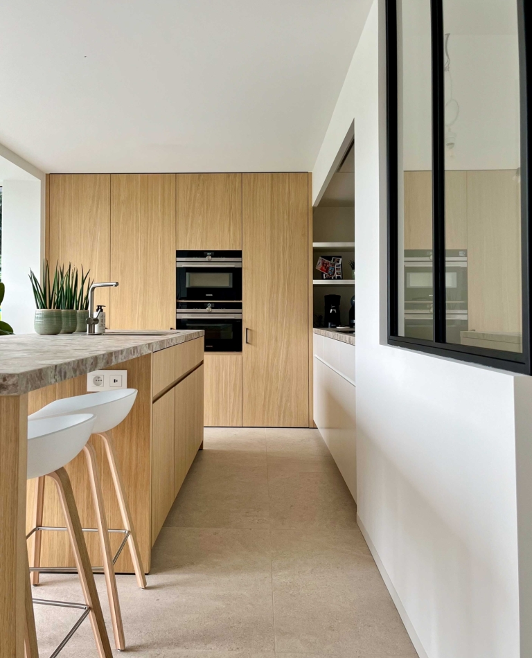 Moderne lichte keuken met natuursteen werkblad, warme houtfineer kasten, grote ramen en strakke baropstelling – maatwerk interieurontwerp door Abstrakt Interieur in België.”