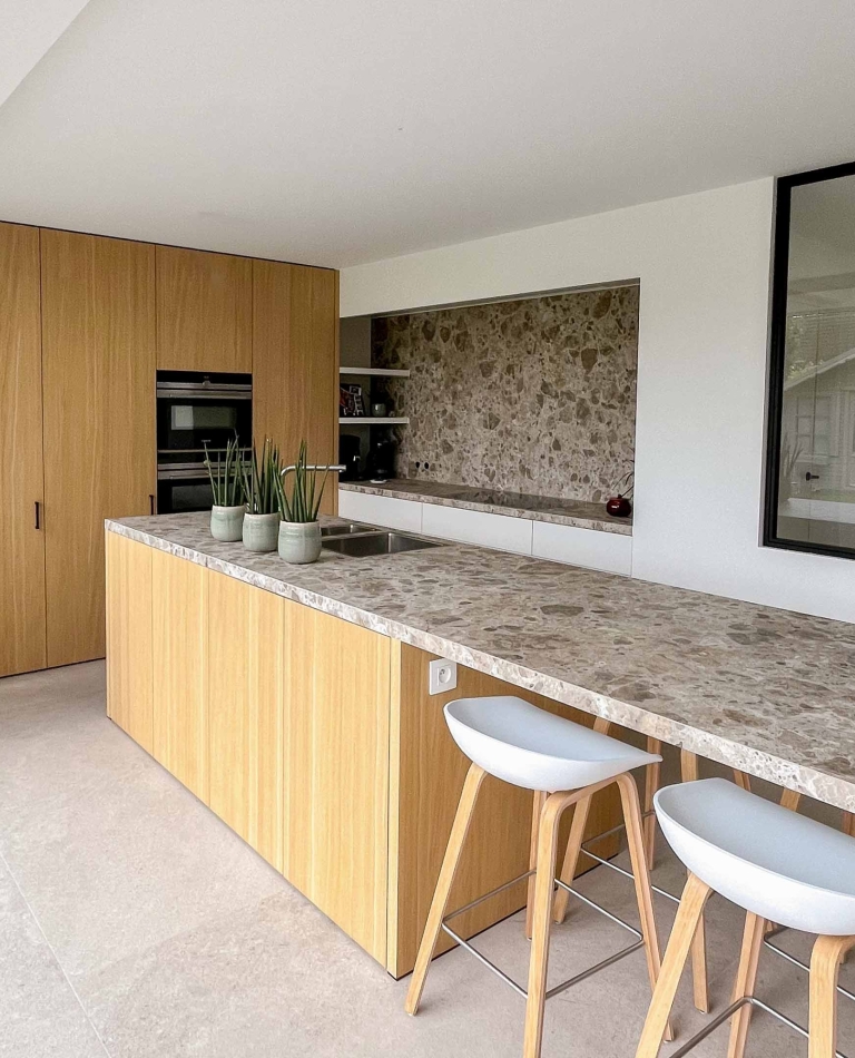 Moderne maatwerkkeuken met eiken kastenwand, groot keukeneiland in natuursteen en witte fronten, ontworpen door Abstrakt Interieur in West-Vlaanderen.