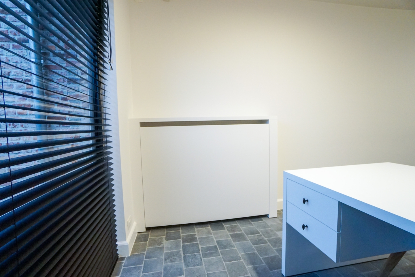 Functioneel maatwerk interieur met witte inbouwkasten, houten legplanken en een geïntegreerd bureau. De doorgang naar extra opbergruimte en de natuursteen vloer versterken het praktische en moderne karakter van deze totaalinrichting in Oostnieuwkerke regio Roeselare West-Vlaanderen