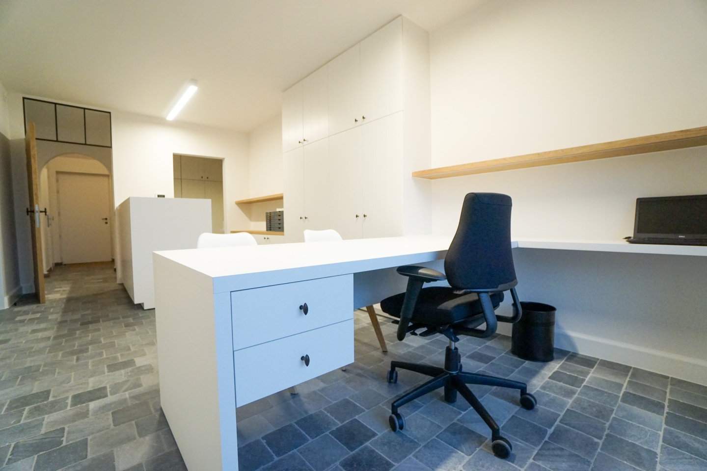 otaalinrichting van een logopediepraktijk in Oostnieuwkerke met maatwerk inbouwkasten, een strak wit bureaublad en houten wandplank. De doorgang met extra opbergruimte en de moderne vloer zorgen voor een uitgepuurd, functioneel geheel