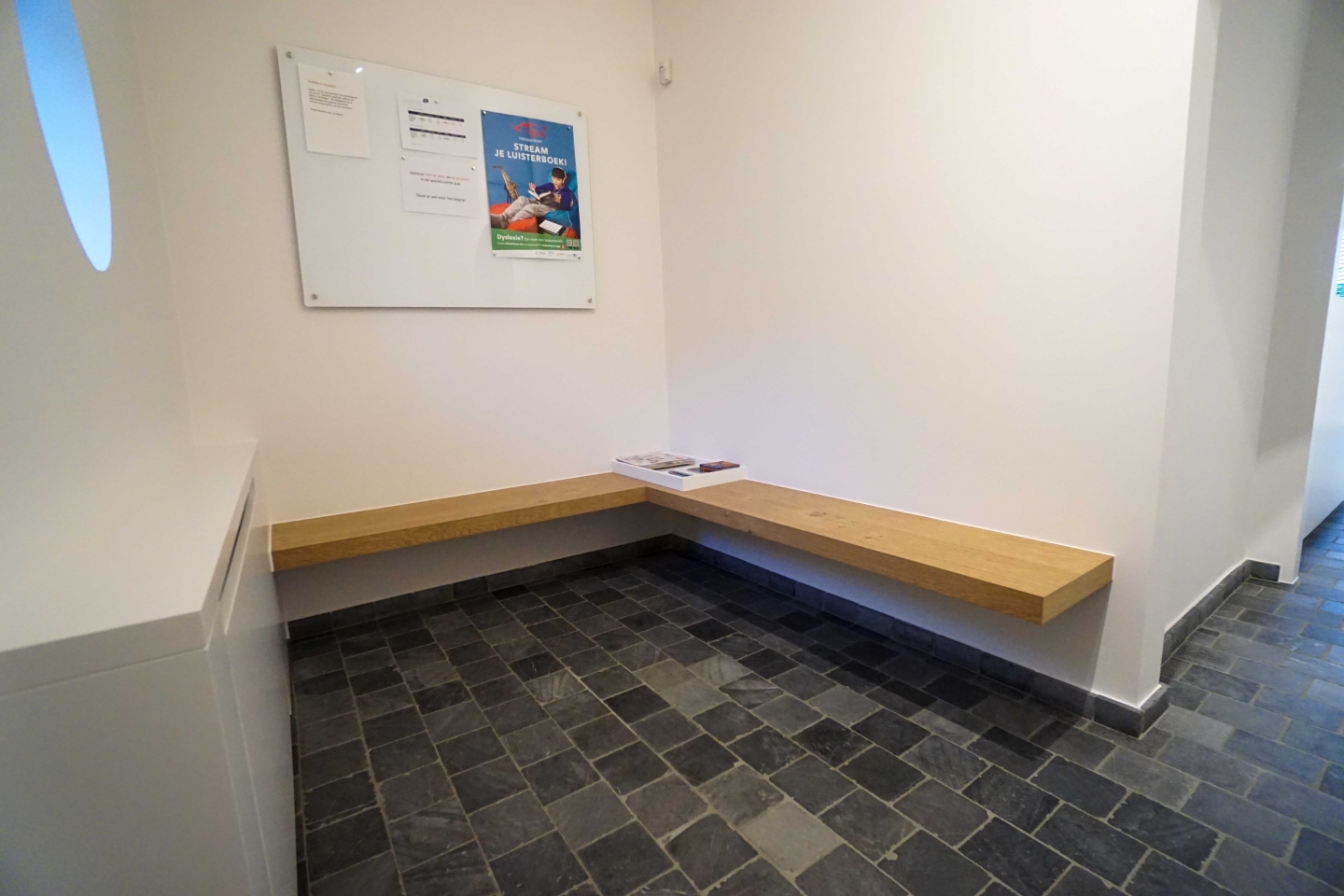Wachtzone in een logopediepraktijk in Oostnieuwkerke met een zwevende L-vormige houten zitbank op maat, natuursteenlook vloer en een wit infobord aan de wand. Onderdeel van een maatwerk interieur en totaalinrichting, ontworpen voor een rustige en overzichtelijke ontvangst