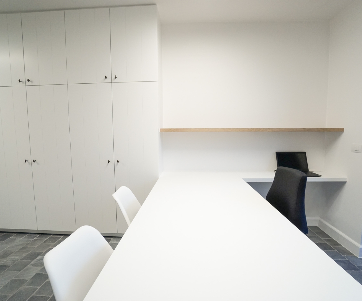 Moderne totaalinrichting van een logopediepraktijk met een volledig op maat gemaakte kastenwand, groot wit consultatiebureau en een aparte werkhoek met zwevend bureaublad. Afgewerkt met een houten accentplank.