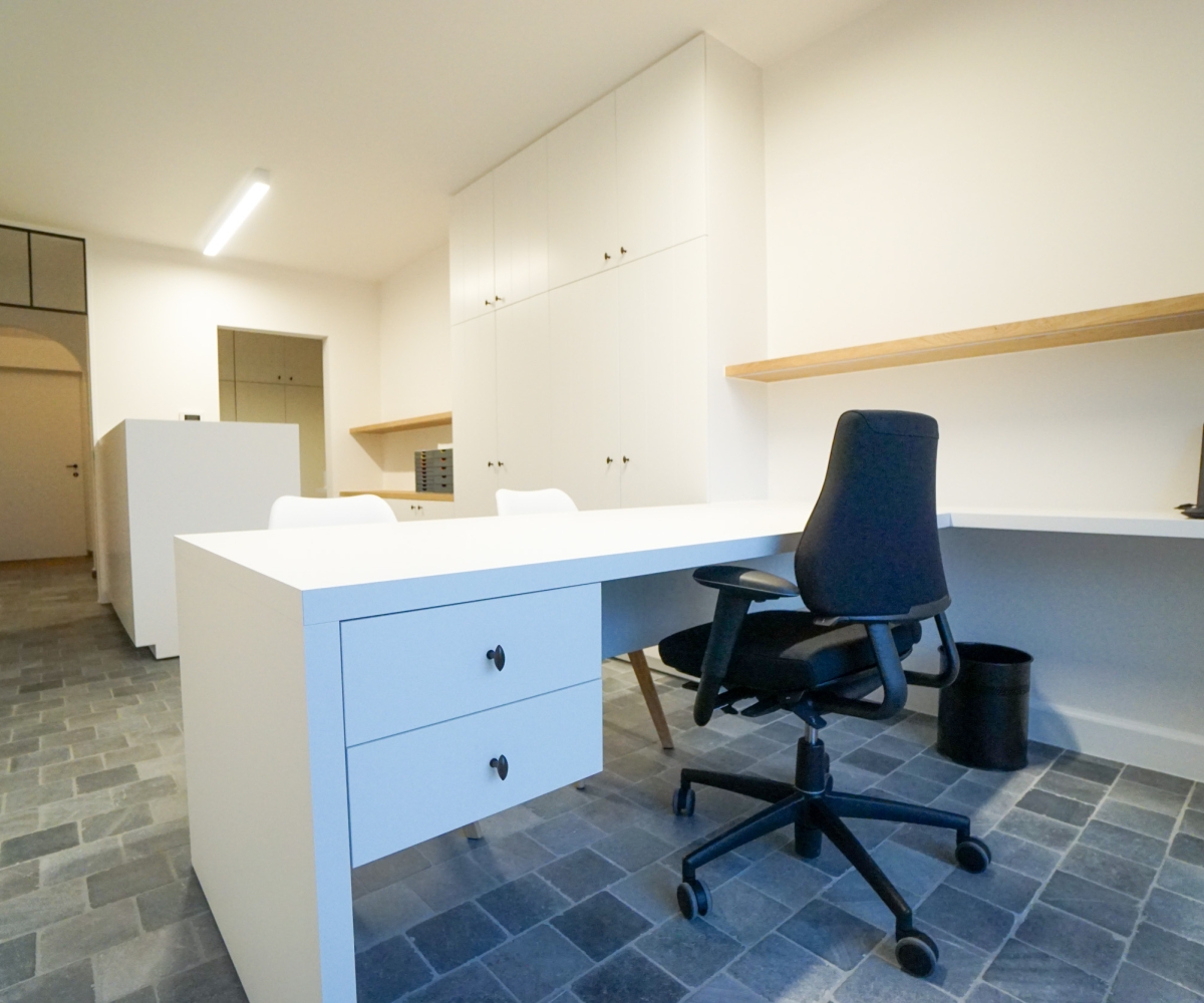 Maatwerk interieur van een praktijkruimte in Oostnieuwkerke met een groot wit bureau op maat, geïntegreerde lades, hoge witte inbouwkasten met verticale belijning en een zwevende houten wandplank. De natuursteenlook vloer, ergonomische bureaustoel en doordachte indeling vormen een moderne en functionele totaalinrichting.”