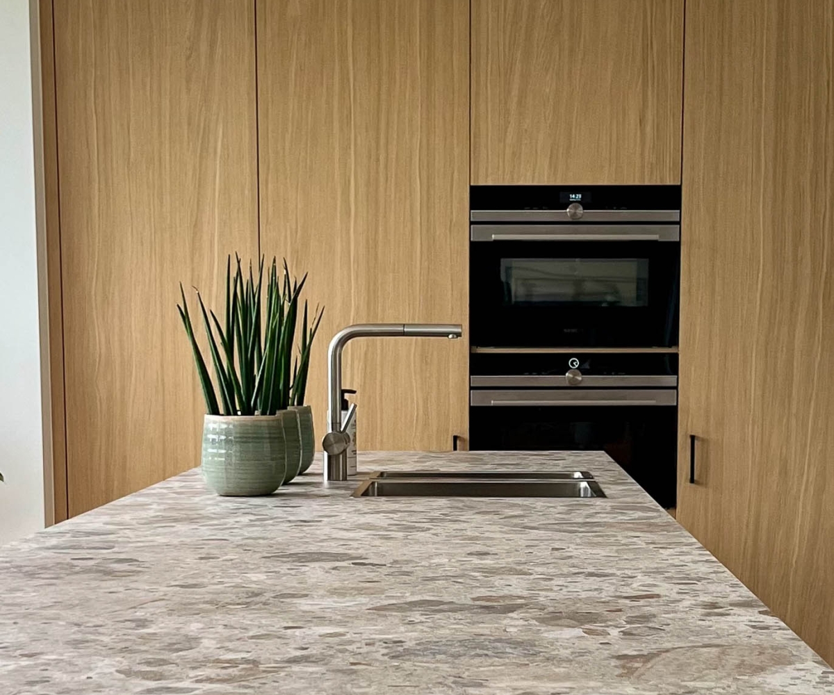 Moderne keuken met natuursteen werkblad, hoge kasten in licht eikenhout en strakke witte onderkasten, met uitzicht op de tuin – kwaliteitsvol maatwerkinterieur door Abstrakt Interieur.”